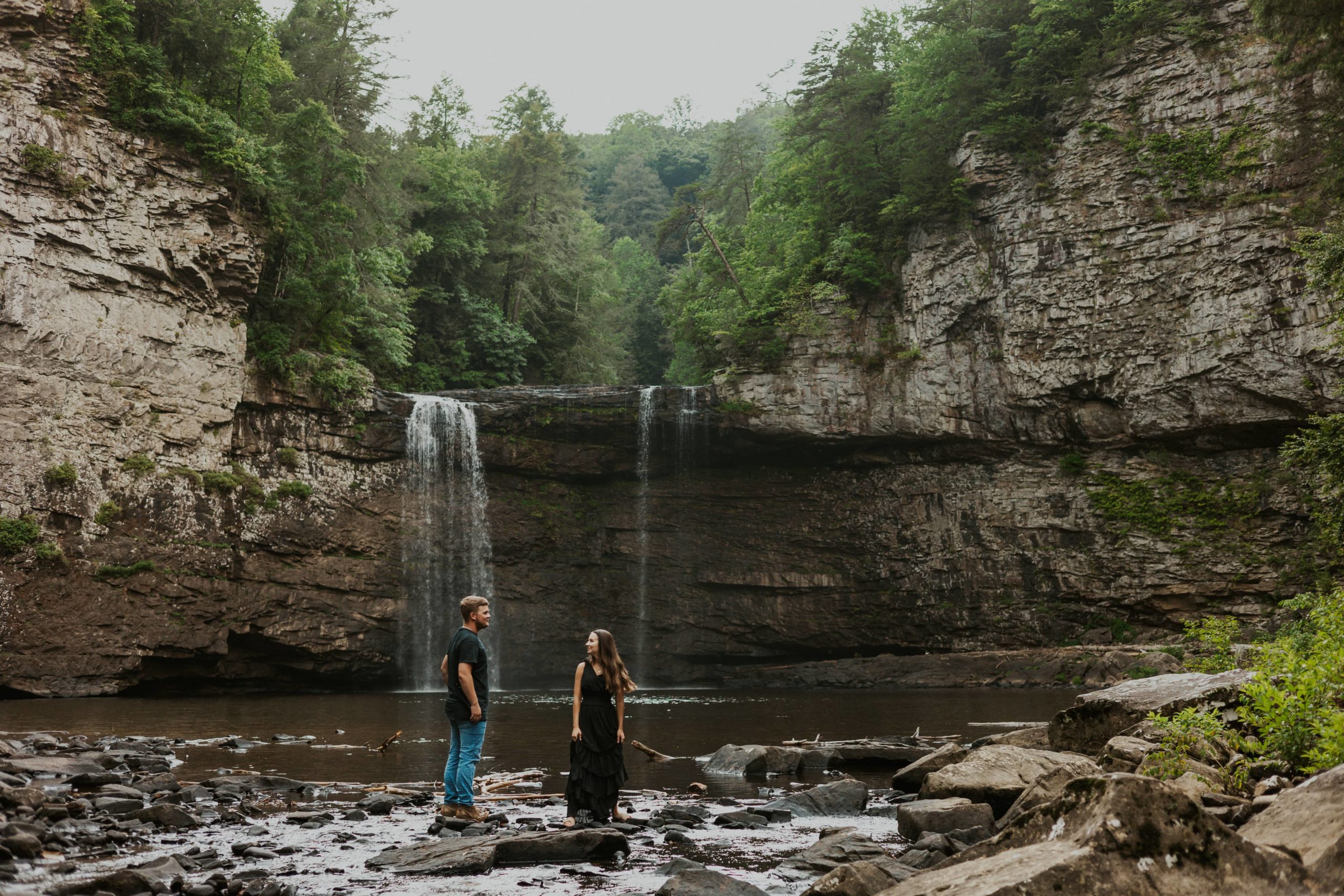 Sydnie and Austin walking across the rocks underneath a waterfall at Fall Creek Falls for their engagement photos.