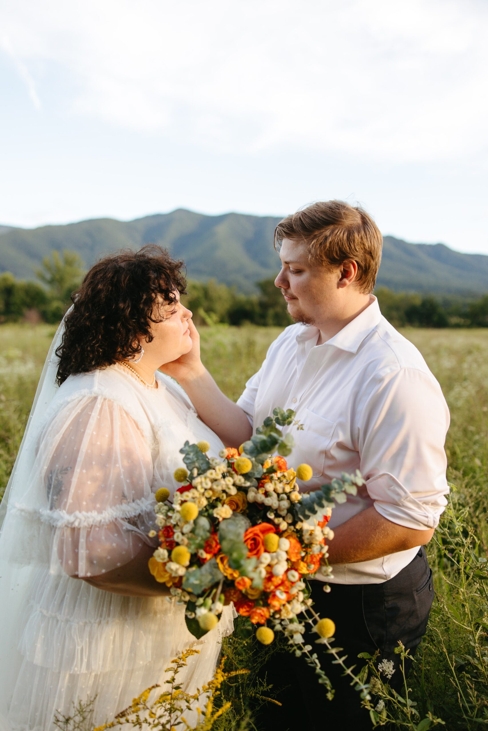 A groom touches his bride's face in a field in the Smoky Mountains.